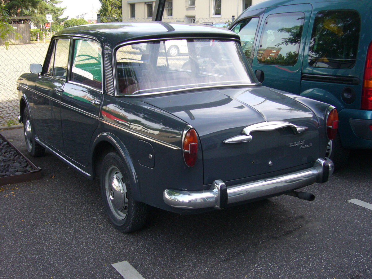 Heckansicht eines Fiat 1100D, wie er von 1962 bis 1966 vom Band lief. Der  Nuova Millecento  wurde bereits 1953 vorgestellt und in etlichen Serien permanent modifiziert. Im April 1962 schickten die Turiner den 1100D ins Rennen. Das D steht hier aber nicht für Diesel, sondern für DERIVAZIONE (Ableitung), da die Karosserie nach einem umfangreichen  Facelifting  auch motorentechnisch aufgerüstet wurde. Für den deutschen und Benelux-Markt wurden die Fahrzeuge (nur die Limousinen) bei NSU-Fiat in Heilbronn montiert. Der Vierzylinderreihenmotor hatte nun einen Hubraum von 1221 cm³ und leistete nun 50 PS. Besucherparkplatz der Düsseldorfer Classic Remise am 15.08.2021.