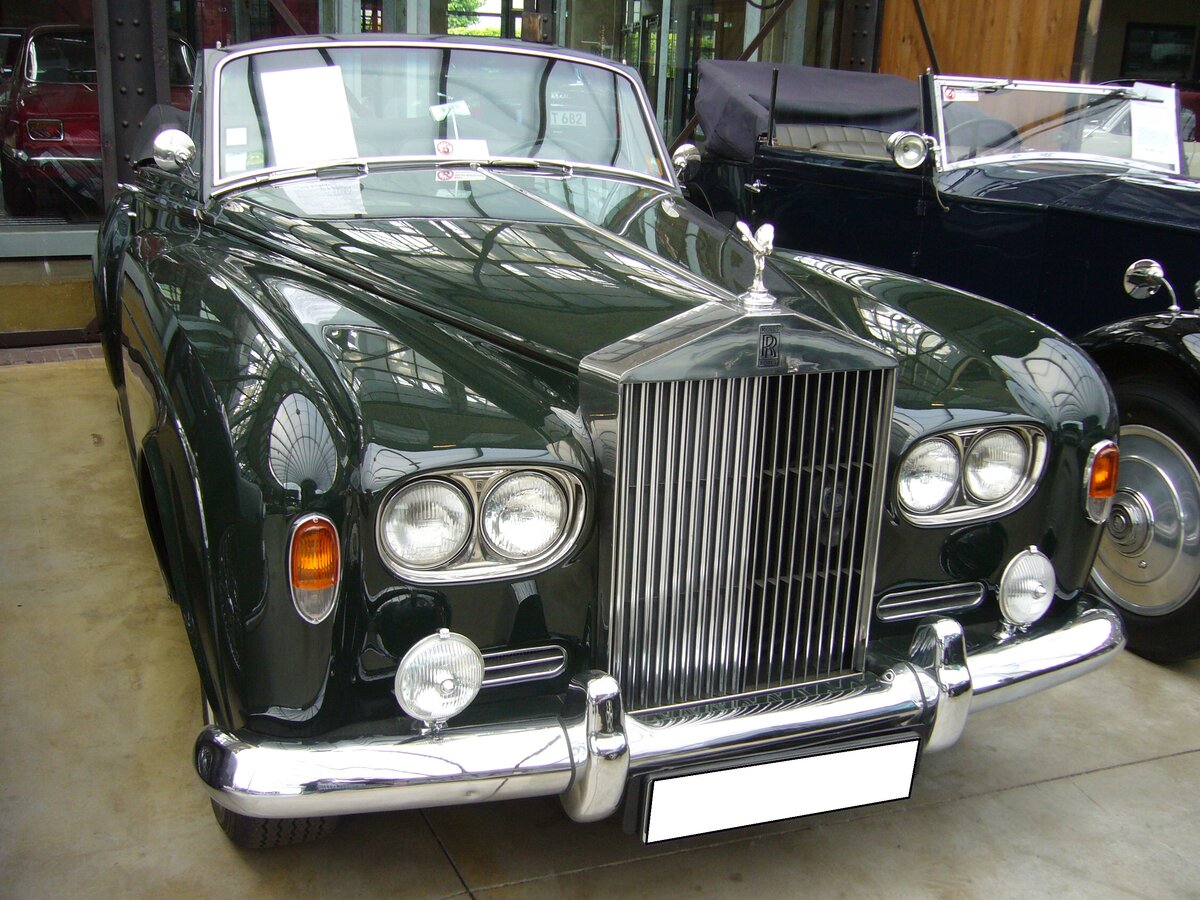 Rolls Royce Silver Cloud III Convertible aus dem Jahr 1964. Produziert wurde dieses Modell t von 1962 bis 1965. Als der Silver Cloud III im Jahr 1962 vorgestellt wurde, fiel dem Fachpublikum als erstes der Wechsel zu Doppelscheinwerfern und eine niedriger geführte Linie der vorderen Kotflügel und der Motorhaube auf. Zum ersten Mal konnte auch das Chassis des Rolls-Royce für den Aufbau einer Coupé- oder Cabriolet-Varianten bestellt werden, die bis dato exklusiv dem fast baugleichen Bentley Continental vorbehalten war. Der V8-Motor hat einen Hubraum von 6230 cm³ und leistet 187 PS. Dieser, im Farbton velvet green lackierte Silver Cloud III wurde 1964 gebaut und ist einer von nur 25 produzierten, linksgelenkten Silver Cloud III Convertible. Ein Vorbesitzer dieses Fahrzeuges ist der US-amerikanische Schauspieler Nicolas Cage. Classic Remise Düsseldorf am 26.05.2022.