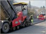 Strassenbauarbeiten in Wiltz. Whrend der LKW den Asphaltfertiger mit Aspahlt befllt und der Zug im Schritttempo weiterrollt wird der Asphalt von dem Asphaltfertiger gleichmssig auf dem Strassenabschnitt verteilt.  13.04.2012