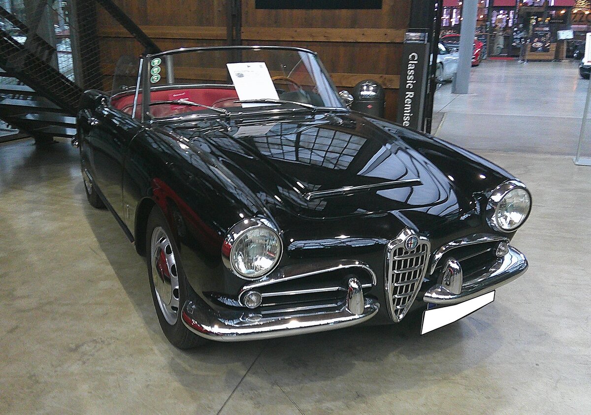 Alfa Romeo Giulia Spider der Baureihe 101. Im Juni 1962 brachte Alfa Romeo die Giulia Spider auf den Markt. Das Modell entsprach optisch fast genau dem Vorgängermodell Giulietta Spider und wurde ebenfalls bei der Carrozzeria Pininfarina montiert. Optisch kann man die Giulia Spider von der Giulietta Spider am besten an der  Lufthutze  auf der Motorhaube erkennen. Von 1962 bis 1965 wurden rund 10.350 Giulia Spider montiert. Der Vierzylinderreihenmotor hat einen Hubraum von 1570 cm³ und leistet 91 PS. Das Modell hatte damals schon serienmäßig ein Fünfganggetriebe. Classic Remise Düsseldorf am 15.01.2026.