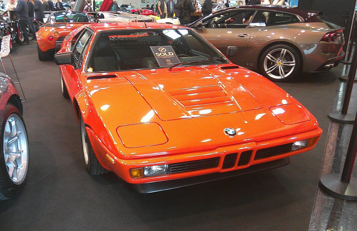 Einer von 460 gebauten BMW E26, uns besser bekannt als BMW M1. Der von 1979 bis 1981 angebotene M1 gehört zu den größten Schöpfungen des Automobilbaus, deren Entstehung spektakulärer war, als sein kommerzieller Misserfolg. Die Technik von BMW, die Karosserie gezeichnet von Giugiaro, das Konzept von Lamborghini und die Produktion bei Baur in Stuttgart. Der als Mittelmotor verbaute Sechszylinderreihenmotor hat einen Hubraum von 3453 cm³ und leistet 277 PS. Das damals DM 113.000,00 teure Auto brachte es auf eine Höchstgeschwindigkeit von 262 km/h. Der gezeigte  M1  dürfte im Farbton liquid orange lackiert sein. Retro Classics in Essen am 09.04.2026.