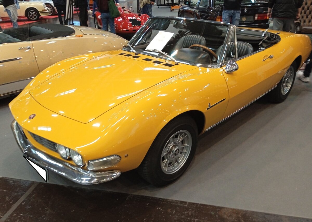 Fiat Dino Spider im Farbton giallo positano, gebaut in den Jahren von 1966 bis 1972. Das Modell Dino war ein  Sondermodell  zum einhundertsten Geburtstag von Giovanni Agnelli (Signore Fiat). Es gab zwei Karosserievarianten: Den Spider gezeichnet von Pininfarina und das Coupe gezeichnet von Bertone. Der Motor war grundsätzlich ein V6-Motor und konnte mit einem Hubraum von 1.988 cm³ und einer Leistung von 160 PS (hauptsächlich für den heimischen, italienischen, Markt) oder 2.418 cm³ Hubraum mit 180 PS bestellt werden. Die Motoren wurden auch im  Dino  von Ferrari und später im Lancia Stratos verbaut. Die Montage der Fahrzeuge erfolgte in Maranello bei Ferrari. Essen Motor Show am 28.11.2025.