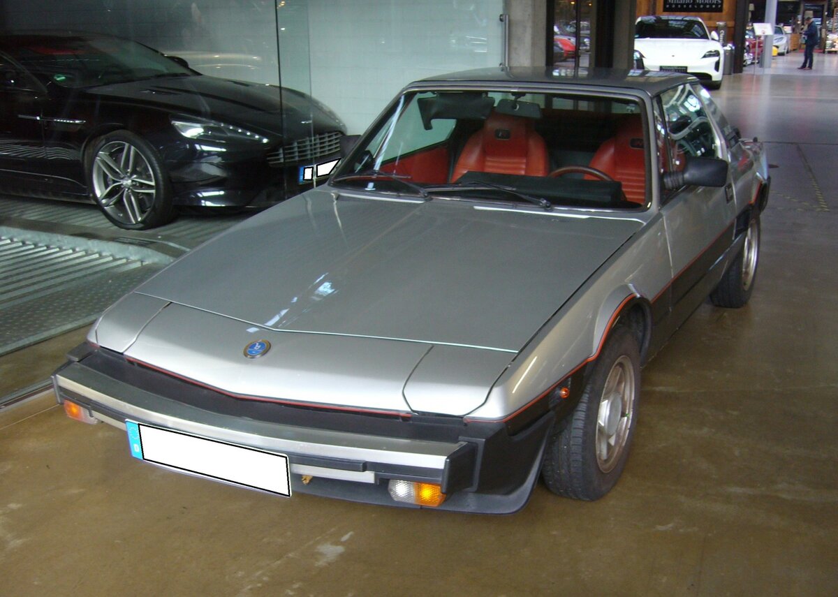 Fiat X1/9 fivespeed im Farbton silbermetallic 656, produziert von 1978 bis 1990. Der X1/9 wurde bereits im Frühjahr 1972 vorgestellt. Er basierte auf dem  Millionenseller  Fiat 128. Bei seiner Vorstellung wurde der Wagen von einem 1.3l Motor mit 75 PS angetrieben. Ab 1978 wurde der 1.5l Motor des Ritmo mit 85 PS verbaut. Seit diesem Zeitpunkt erhielt das Auto den Zusatz five speed. Ab dem Jahr 1982 wurde der keilförmige, zweisitzige Sportwagen bei Bertone produziert. Classic Remise Düsseldorf am 30.11.2023.