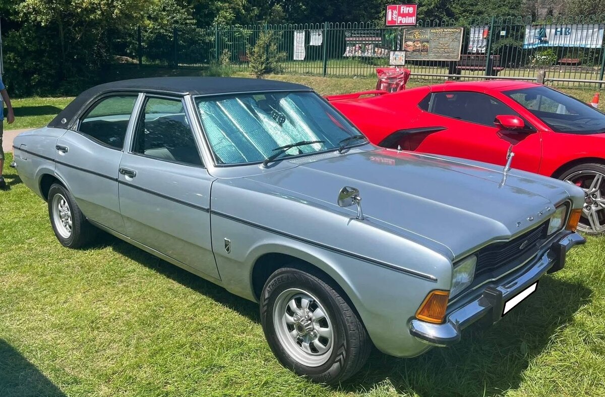 Ford Cortina MK3 2000E im Farbton silver fox und der Karosserieversion Limousine viert�rig. Der  Cortina MK3 , gebaut von 1970 bis 1976 ist im Grunde die britische Version des deutschen Ford Taunus TC. �u�erlich und technisch unterschieden sich die Modelle jedoch etwas. Der MK3 ersetzte das Vorg�ngermodell Cortina MK2, als auch den gr��eren Corsair. Um Kosten zu sparen, teilte er sich die Plattform mit dem deutschen Taunus. Daher die Abk�rzung TC f�r Taunus & Cortina. Im Vergleich zum Vorg�nger war er breiter und hatte einen l�ngeren Radstand, wodurch der Innenraum wesentlich ger�umiger ausfiel. Der Vierzylinderreihenmotor hat einen Hubraum von 1993 cm� und leistet 99 PS. 