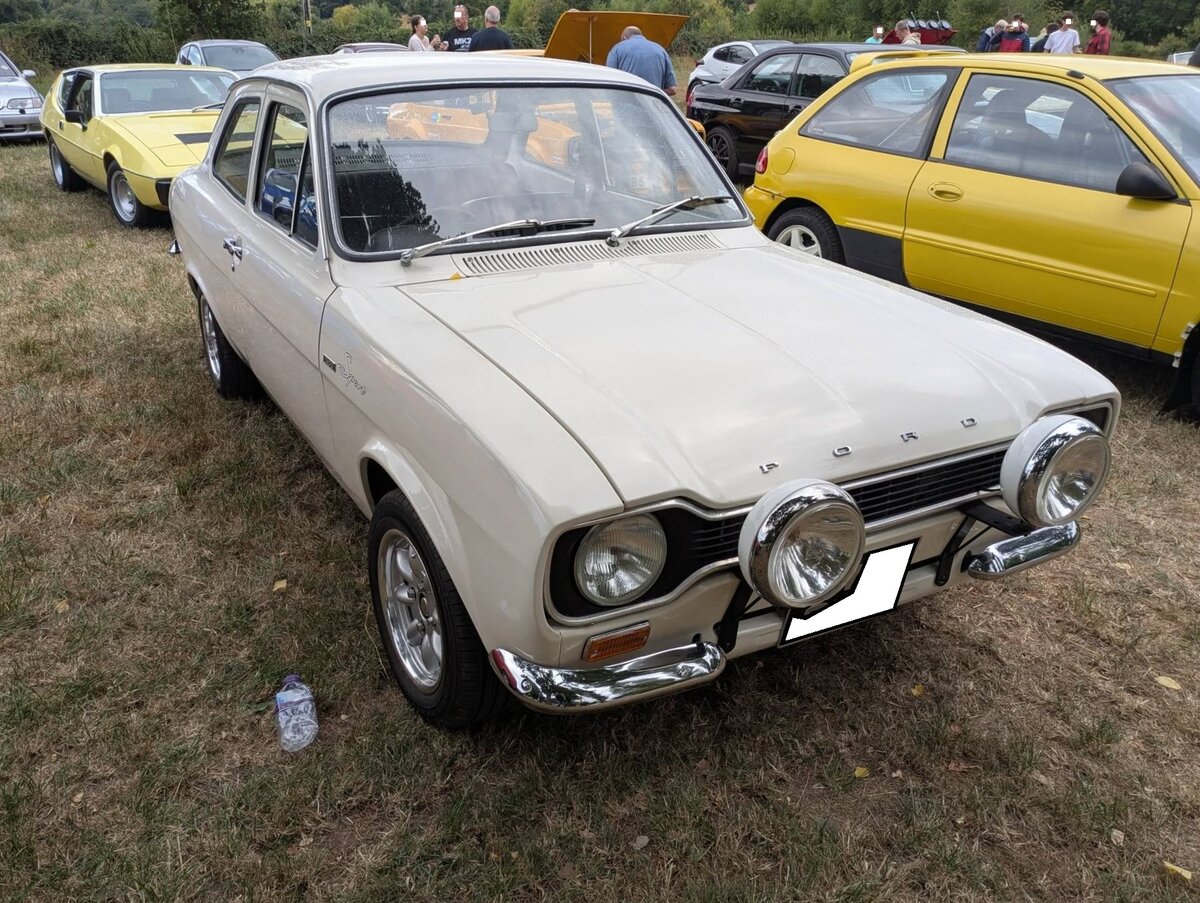 Ford Escort MK1 1300 Sport aus dem Jahr 1971 im Farbton diamond white. In Deutschland hie� der Farbton diamant wei�. Im August 1968 pr�sentierte man unter dem Motto:  Familienwagen und sportlicher Flitzer  die erste Escort-Generation. Ihr schmaler K�hlergrill, der in die Scheinwerfer m�ndet, bringt ihm sp�ter den Spitznamen  Hundeknochen  ein. Der in England entwickelte Nachfolger des legend�ren  Anglia  wurde der Escort sp�ter auch in den Ford-Werken Genk/Belgien und Saarlouis/Deutschland gebaut. Der gezeigte 1300 Sport kam im Jahr 1971 auf den Markt und war im Grunde eine g�nstigere, abgespeckte, Variante des bereits seit 1969 recht beliebten 1300 GT. Er hatte die sportlichen Technikkomponenten und das Fahrwerk des 1300 GT, bot jedoch einen einfacheren Innenraum. Der Vierzylinderreihenmotor namens Kent-Crossflow hat einen Hubraum von 1298 cm� und leistet, je nach Vergaserbest�ckung, zwischen 57 PS und 72 PS. Treffen des Chainbridge classic car club auf der Wiese vor einem Pub in Chainbridge/Wales im Juli 2025.