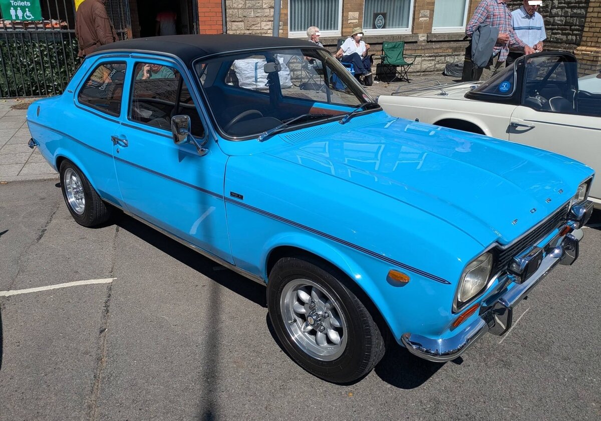 Ford MK1 Escort 1300E. Dem zunehmenden Wandel auf dem europ�ischen Markt Ende der 1960´er Jahre kommt Ford gleich mit zwei Neuentwicklungen entgegen. Im August 1968 pr�sentierte man unter dem Motto:  Familienwagen und sportlicher Flitzer  die erste Escort-Generation. Ihr schmaler K�hlergrill, der in die Scheinwerfer m�ndet, bringt ihm sp�ter den Spitznamen  Hundeknochen  ein. Als in England entwickelter Nachfolger f�r den Anglia wurde der Escort sp�ter auch in den Ford-Werken Genk/Belgien und Saarlouis/Deutschland gebaut. Europaweit war er Fords bisher erfolgreichstes Modell, in Deutschland ersetzte er den gl�cklosen P6, konnte sich jedoch gegen die Konkurrenz aus R�sselsheim kaum behaupten. Das lag zum einen an seiner pummeligen Form und der simplen Technik, sowie der schlechten Stra�enlage aufgrund der hinteren Blattfedern. Auch eine merkw�rdige Modellpolitik trug ihren Teil dazu bei: So waren zuerst weder heizbare Heckscheibe noch Schiebedach lieferbar. In England war der Escort ab Jahresanfang 1968 erh�ltlich und verkaufte sich gl�nzend. Hier haben wir einen rechts gelenkten, zweit�rigen, Escort MK1 im Farbton olympic blue. Der Vierzylinderreihenmotor hat einen Hubraum 1298 cm� und leistet im 1300E 72 PS. Brynmawr Classic Car Show/South Wales am 26.07.2026.
