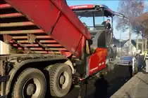 Der Asphalt wird vom Kipper in den Schlund des Asphaltfertigeres geschüttet, um damit über die ganze Straßen breite gleichmäßig verteilt zu werden.  (11.2018)